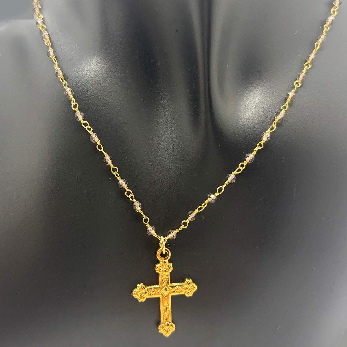 Fotografía real del Collar Bruma sobre busto negro, con cuentas de cuarzo ahumado, cadena dorada tipo rosario y colgante de cruz dorada.