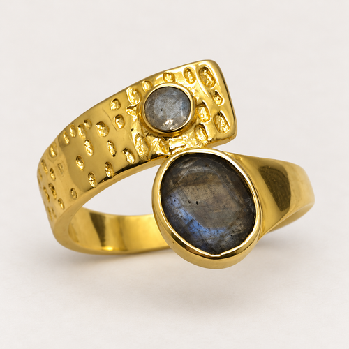 Anillo Eclipse de tono dorado con una piedra ovalada y otra redonda en tonos gris azulados, sobre fondo gris claro.