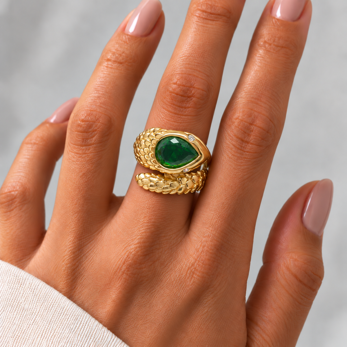 Anillo Serp con diseño de serpiente, acabado dorado y circonita verde, sobre la mano de una modelo con manicura natural.