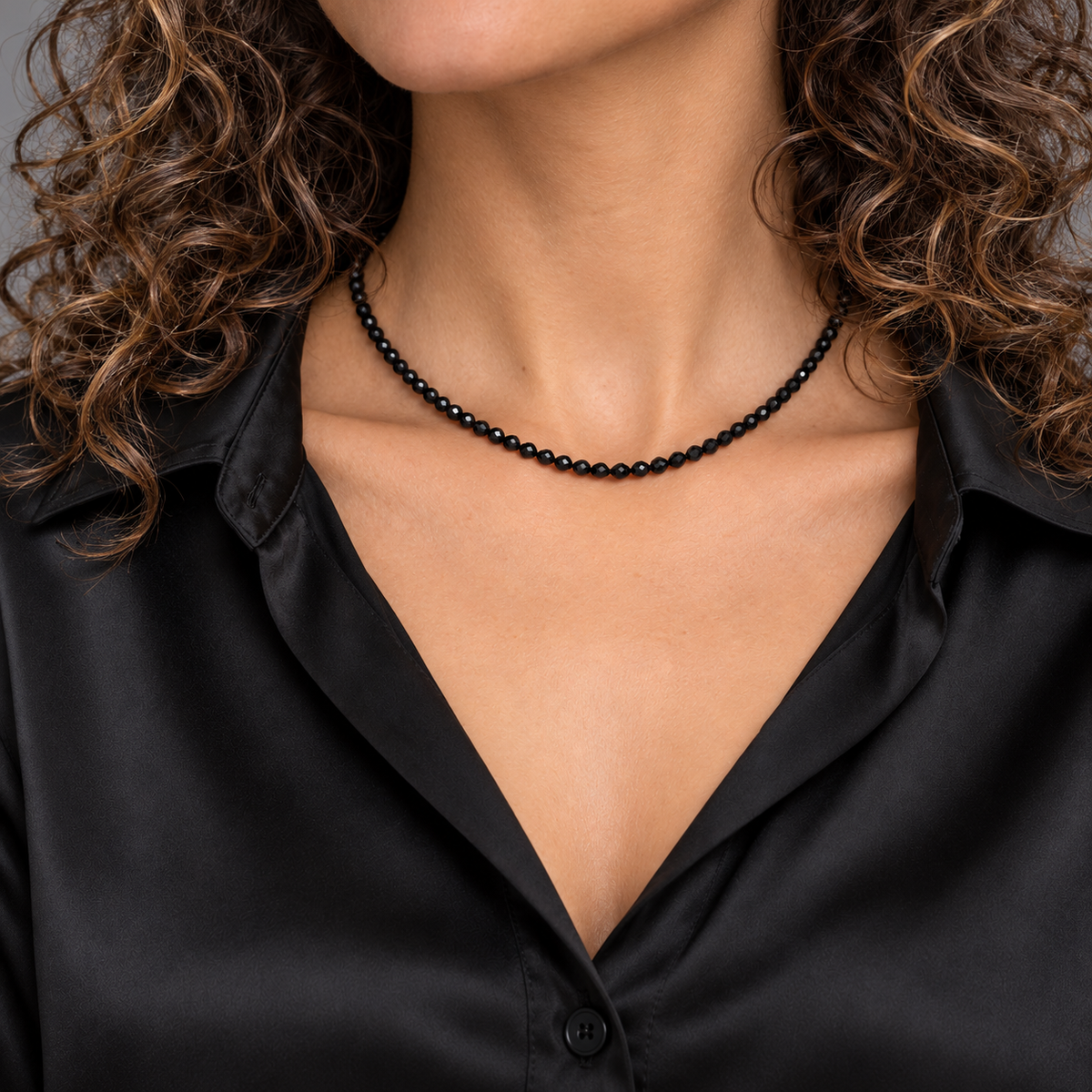 Choker de ónix negro facetado de 3 mm sobre modelo de cabello rizado y blusa de seda negra, encuadrada desde la barbilla.
