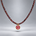 Collar Cap Roig con cuentas de granate y colgante circular de rubí reconstituido rosado en plata chapada en oro, suspendido sobre fondo gris.