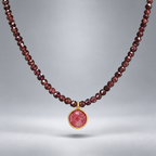 Collar Cap Roig con cuentas de granate y colgante circular de rubí reconstituido rosado en plata chapada en oro, suspendido sobre fondo gris.