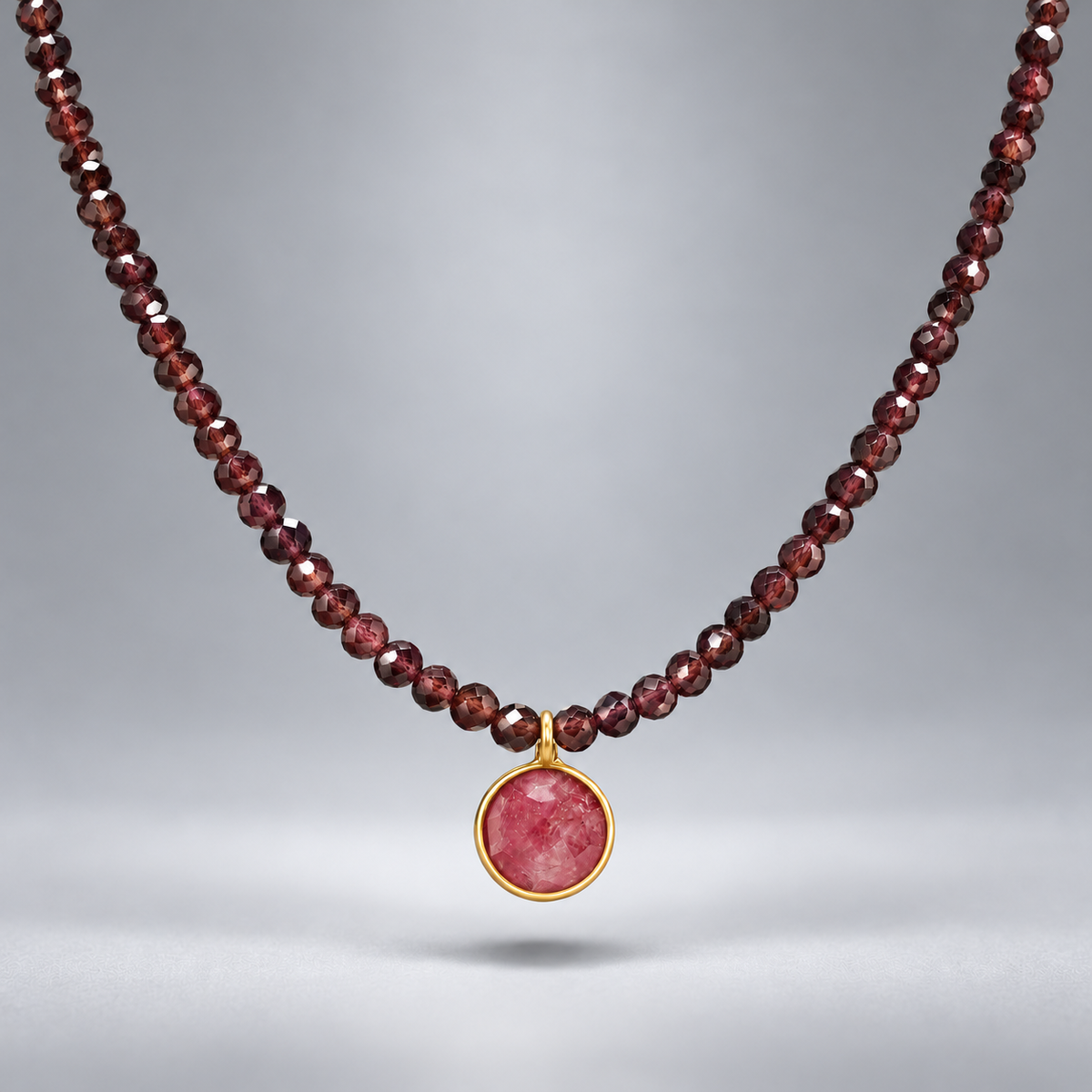 Collar Cap Roig con cuentas de granate y colgante circular de rubí reconstituido rosado en plata chapada en oro, suspendido sobre fondo gris.