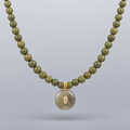 Collar Sa Riera de jade verde oliva, con colgante circular y dos piezas redondas frontales de acero chapado en oro, suspendido sobre fondo gris claro degradado.