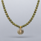 Collar Sa Riera de jade verde oliva, con colgante circular y dos piezas redondas frontales de acero chapado en oro, suspendido sobre fondo gris claro degradado.