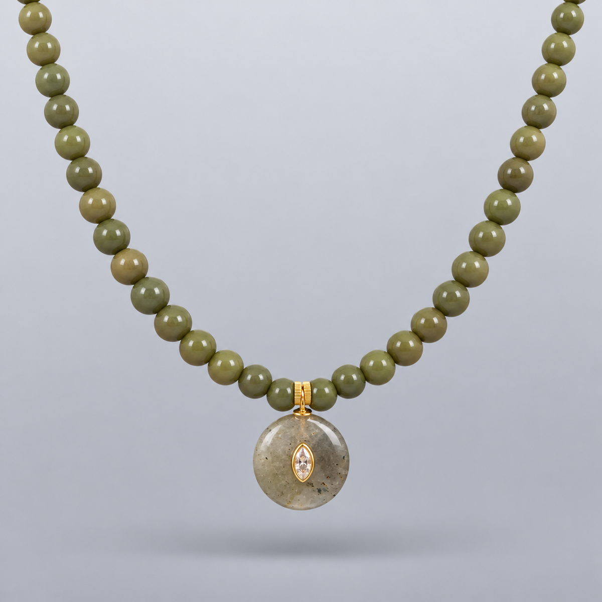 Collar Sa Riera de jade verde oliva, con colgante circular y dos piezas redondas frontales de acero chapado en oro, suspendido sobre fondo gris claro degradado.